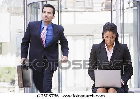 Stock Photograph - Businesswoman outdoors in front of building using laptop with businessman in background (high key/blur). Fotosearch