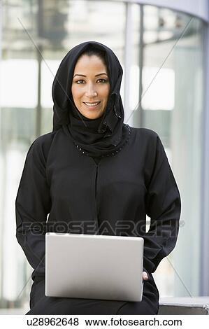 Stock Photo - Businesswoman sitting outdoors with laptop by building smiling (selective focus). Fotosearch