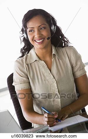 Stock Image - Businesswoman wearing headset in office by laptop smiling (high key/selective focus). Fotosearch