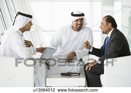 Stock Image - Three businessmen indoors with a laptop talking and smiling (high key/selective focus). Fotosearch