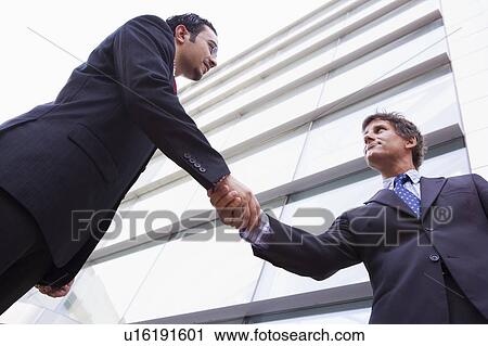 Stock Image - Two businessmen outdoors by building shaking hands (high key/selective focus). Fotosearch