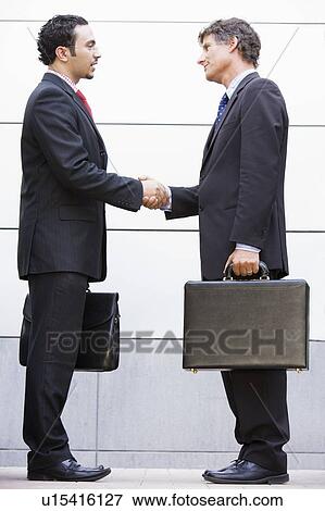 Stock Photo - Two businessmen shaking hands outdoors holding briefcases and smiling. Fotosearch