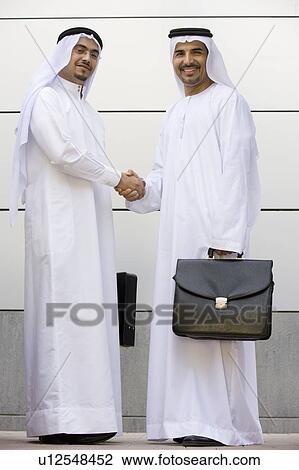 Stock Image - Two businessmen standing outdoors with briefcases shaking hands and smiling. Fotosearch
