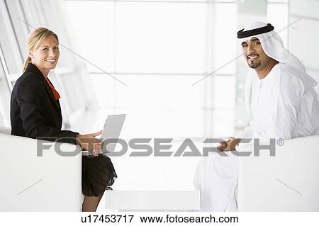 Stock Photo - Two businesspeople indoors with a laptop smiling (high key/selective focus). Fotosearch