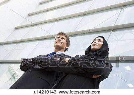 Stock Image - Two businesspeople standing outdoors by building smiling (selective focus). Fotosearch
