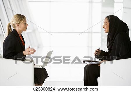Two businesswomen indoors with a laptop talking and smiling (high key/selective focus) View Large Photo Image Picture - Two businesswomen indoors with a laptop talking and smiling (high key/selective focus). Fotosearch