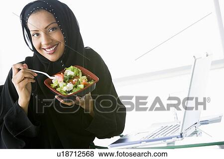 Stock Photo - Woman eating salad near laptop smiling (high key/selective focus). Fotosearch