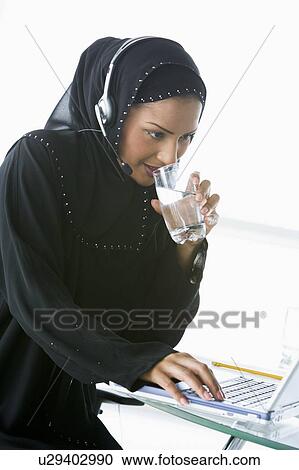 Stock Image - Woman wearing headset with laptop drinking water and smiling (high key/selective focus). Fotosearch