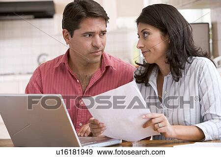 Stock Photo - Couple in kitchen with paperwork using laptop looking unhappy. Fotosearch