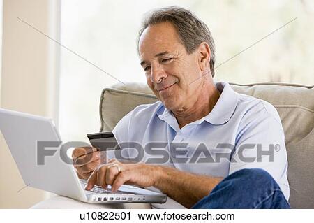 Stock Image - Man in living room with laptop and credit card smiling. Fotosearch