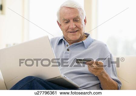 Stock Image - Man in living room with laptop and credit card smiling. Fotosearch