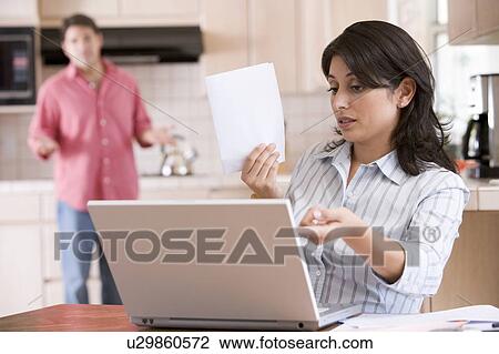 Stock Image - Woman in kitchen with paperwork using laptop with man in background. Fotosearch