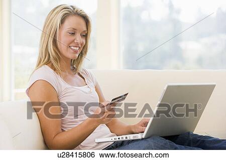 Stock Photograph - Woman in living room using laptop and holding credit card smiling. Fotosearch