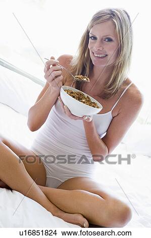 Picture - Woman sitting on bed eating cereal smiling. Fotosearch