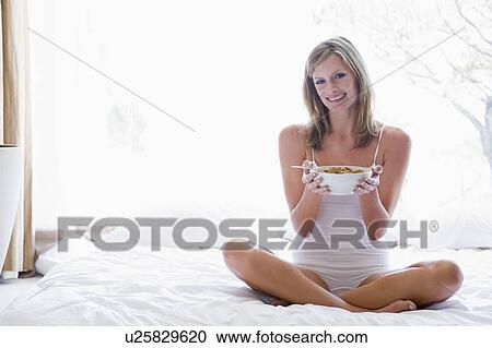 Stock Image - Woman sitting on bed eating cereal smiling. Fotosearch
