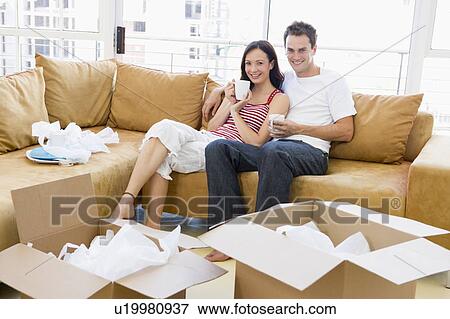 Stock Photo - Couple relaxing with coffee by boxes in new home smiling. Fotosearch