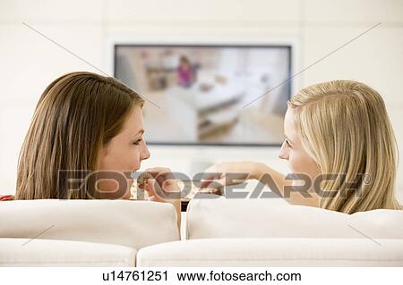 Stock Image - Two women in living room watching television eating chocolates smiling. Fotosearch