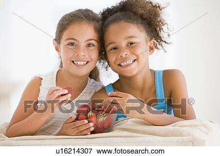 Stock Photo - Two young girls eating strawberries in living room smiling. Fotosearch