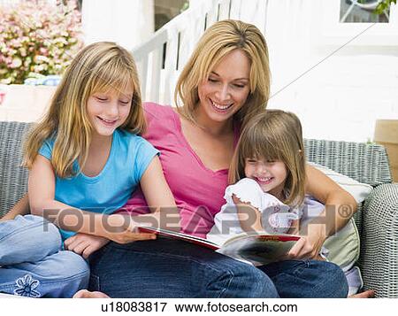 Stock Photo - Woman and two young girls sitting on patio reading book smiling. Fotosearch