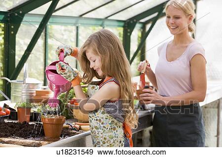 Young girl in greenhouse watering plant with woman holding pot smiling View Large Photo Image Stock Photo - Young girl in greenhouse watering plant with woman holding pot smiling. Fotosearch