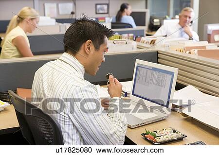 Stock Image - Businessman in cubicle at laptop eating sushi. Fotosearch