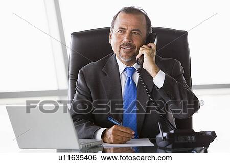 Stock Photograph - Businessman in office on telephone by laptop smiling (high key/selective focus). Fotosearch