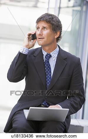 Businessman sitting in office lobby with laptop using cellular phone View Large Photo Image Stock Photograph - Businessman sitting in office lobby with laptop using cellular phone. Fotosearch