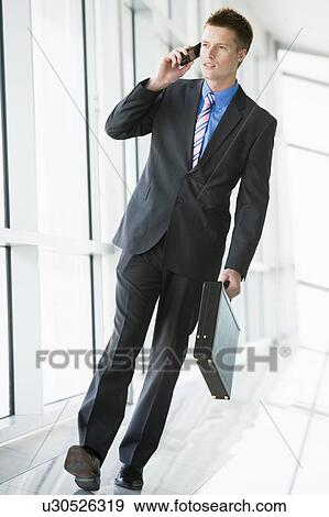 Businessman walking in corridor using cellular phone View Large Photo Image Stock Photo - Businessman walking in corridor using cellular phone. Fotosearch