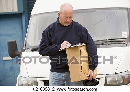 Stock Image - Deliveryperson standing with van writing in clipboard holding box smiling. Fotosearch