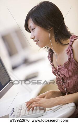 Stock Photograph - Woman in computer room listening to MP3 player while typing and smiling. Fotosearch