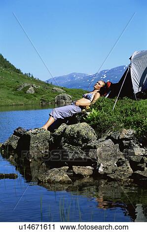 Stock Image - Woman outdoors at campsite sleeping on large rocks by lake. Fotosearch