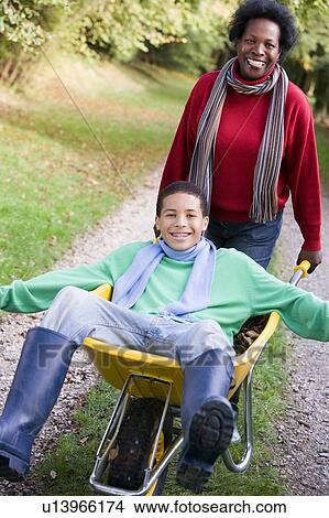 Picture - Mother outdoors pushing son in wheelbarrow and smiling (selective focus). Fotosearch