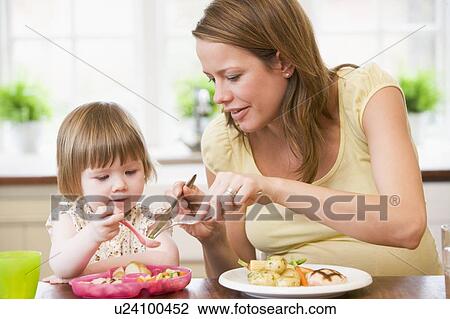 Stock Image - Pregnant mother in kitchen eating chicken and vegetables helping daughter eat. Fotosearch