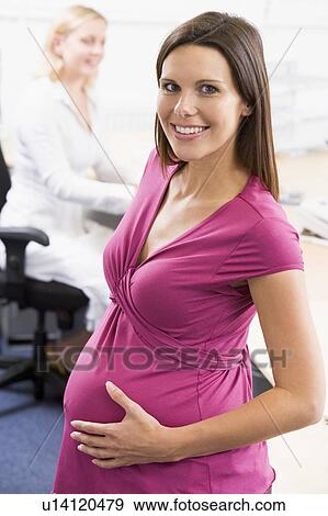 Pregnant woman at work holding belly smiling with coworker in background View Large Photo Image Stock Photo - Pregnant woman at work holding belly smiling with coworker in background. Fotosearch