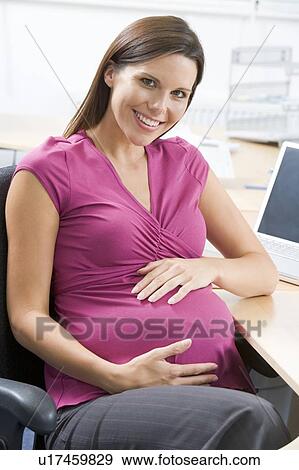 Stock Photo - Pregnant woman at work holding belly and smiling. Fotosearch