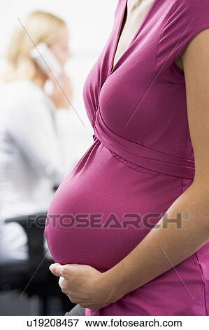 Stock Photo - Pregnant woman at work holding belly with coworker in background. Fotosearch