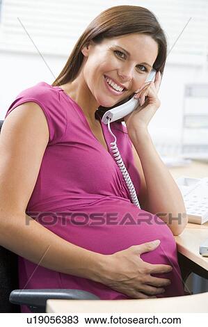 Stock Image - Pregnant woman at work using telephone smiling. Fotosearch