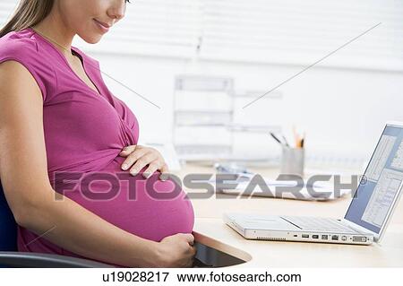 Stock Photo - Pregnant woman at work with laptop holding belly and smiling. Fotosearch