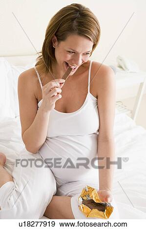 Stock Photo - Pregnant woman in bed eating chocolate smiling. Fotosearch