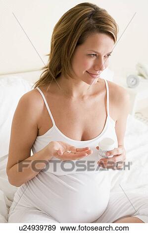 Stock Photo - Pregnant woman in bedroom holding medicine and water smiling. Fotosearch