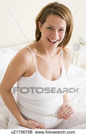 Stock Photograph - Pregnant woman in bedroom measuring belly smiling. Fotosearch