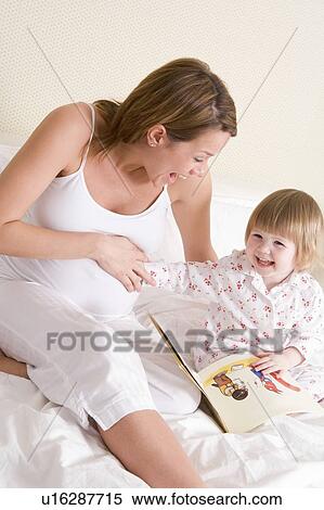 Stock Photography - Pregnant woman in bedroom reading book with daughter laughing. Fotosearch