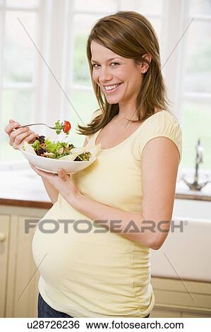 Stock Photograph - Pregnant woman in kitchen eating a salad smiling. Fotosearch