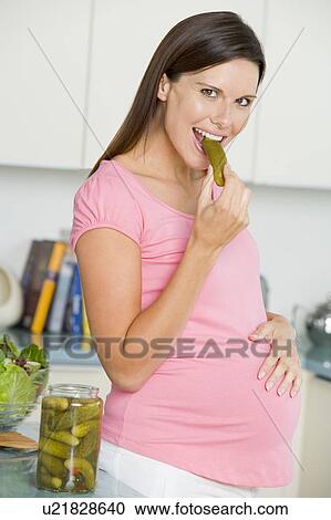 Stock Image - Pregnant woman in kitchen eating pickles and smiling. Fotosearch