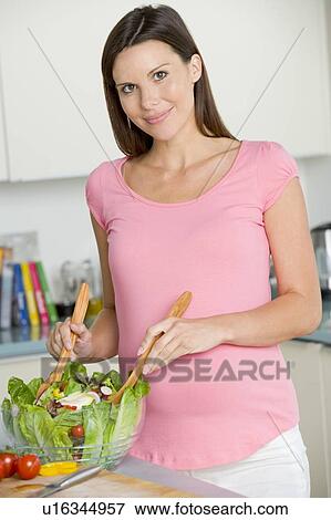 Stock Photo - Pregnant woman in kitchen making a salad and smiling. Fotosearch