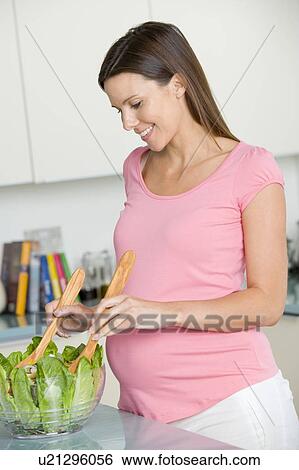 Stock Photograph - Pregnant woman in kitchen making a salad and smiling. Fotosearch