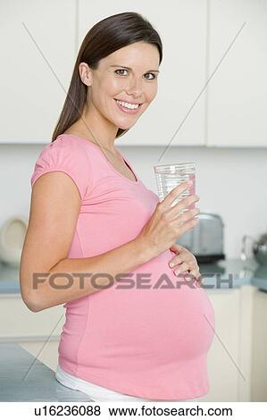 Pregnant woman in kitchen with glass of water smiling View Large Photo Image Stock Photo - Pregnant woman in kitchen with glass of water smiling. Fotosearch