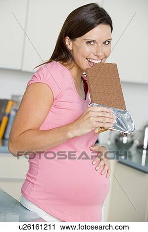 Stock Image - Pregnant woman in kitchen with large chocolate bar smiling. Fotosearch