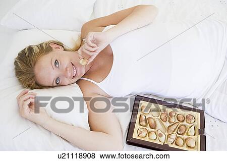 Stock Photo - Pregnant woman lying in bed with chocolates smiling. Fotosearch