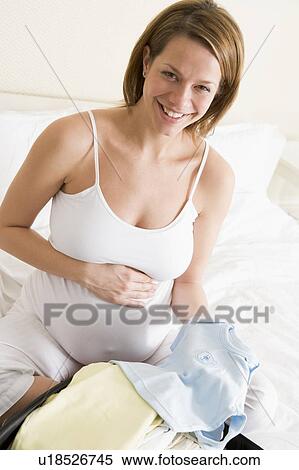 Stock Photography - Pregnant woman packing baby clothing in suitcase smiling. Fotosearch